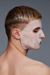 Vertical side view portrait of blonde young man wearing face mask enjoying male skincare against grey background