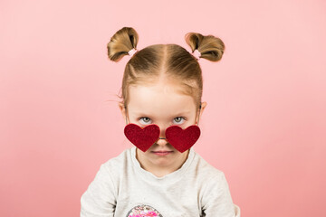 Funny angry little blonde girl in red heart shape sunglasses on pink background. Valentines Day or Mothers Day concept