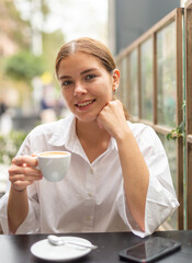 Smiling stylish young woman in white blouse drinking coffee sitting at the cafe
