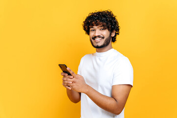 Happy modern attractive Indian or Arabian guy in white t-shirt, uses mobile phone, texting with...
