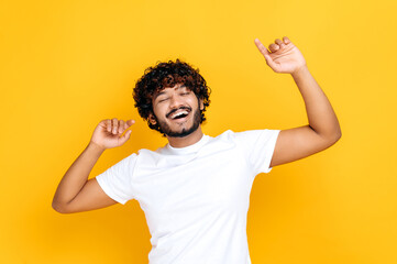 Joy, victory, luck, dancing. Cheerful happy Indian guy in casual white t-shirt, dancing to favorite music, having fun, rejoice in luck, celebrating win, on isolated orange background, smiling