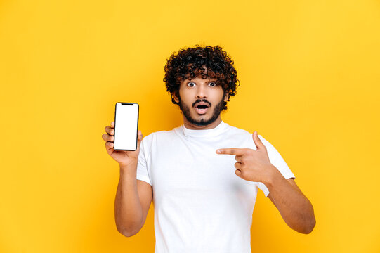 Shocked surprised Indian guy in white t-shirt, holds smartphone with mock-up empty white screen and points finger at it, stands on isolated orange background, shocked looks at camera
