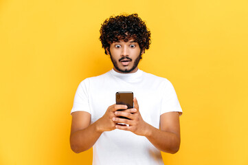 Surprised shocked Indian guy in white t-shirt, uses his smartphone, looks in amazement at screen,...