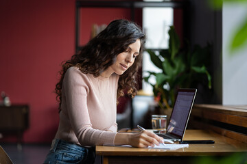 Young concentrated woman auditor sitting in front of laptop and doing paperwork at workplace in office, focused female employee preparing audit report or financial statement. Selective focus
