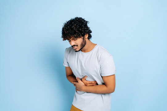 Stomach ache. Indian guy with in casual clothes hunching and holding his belly, suffers from pain in the abdomen or stomach, got poisoned, need treatment, stands on isolated blue background
