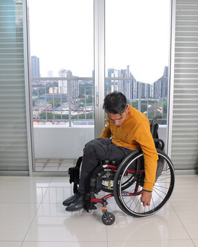 South east Asian malay middle aged disabled man on wheelchair looking sitting in front of balcony window picking up