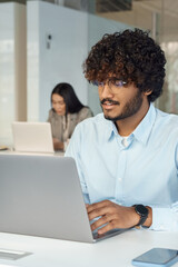 Busy smart young indian man in glasses works on laptop sitting at desk