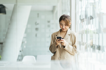 Fototapeta premium Asian woman with smartphone standing against street blurred building background, Fashion business photo of beautiful girl in casual suite with smart phone.