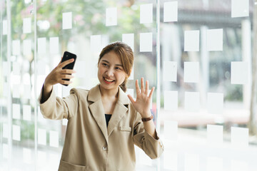 Fototapeta premium Asian business woman waving hand using smartphone app enjoying online virtual chat video call with friends in distance mobile chat virtual meeting, recording stories for social media at home.