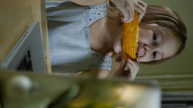portrait, vertical screen: teenager Girl in a dress eats boiled sweet corn and looks into a laptop. slow motion