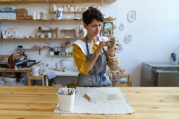 Concentrated woman potter holding paintbrush, painting on clay jug in workshop, working in pottery...