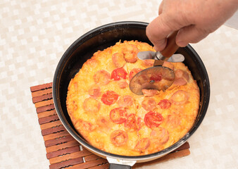 Ready to eat homemade pizza with sausages, tomatoes and cheese being cut with a pizza cutter by senior woman hand. Top view with selective focus. Process of cooking pizza, step 13