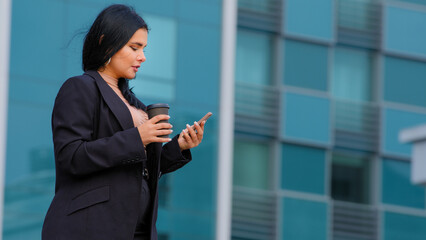 Young hispanic businesswoman walking street background of office building checks email on phone uses mobile app makes purchase online sends text message on smartphone drinking coffee from paper cup