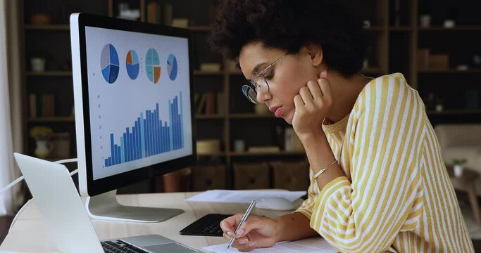 Serious African businesswoman sit at desk holds pen writes, makes market research working with graphs and financial statistics shown on computer monitor, prepare stats report use modern tech concept