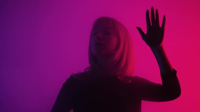Woman emerges from smoke and touches glass with palms. Blonde examines glass and slides her hands over it with interest and surprise. Model poses in dark studio with pink and purple lights. Close up.