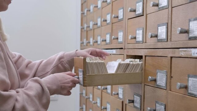 4K. A young caucasian blonde woman is looking for information in a file cabinet. Female student looking for necessary literature in library reference card catalog. Search documents in the card index