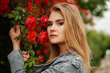 A cute blonde stands near a bush with red roses. Attractive European woman in a denim jacket.