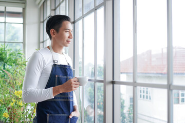 Fototapeta premium Happy young Asian man wearing apron smiling and holding cup of coffee looking out of window after take care of trees in greenhouse at home
