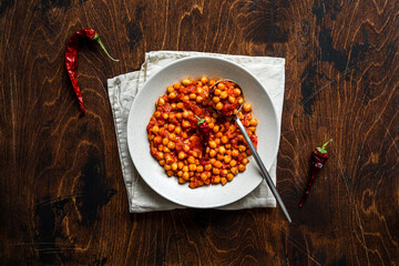 A bowl of stewed cheak-pea with hot tomato sauce on a napkin, on wooden background, overhead shot. A spoon and red chilli pepper.