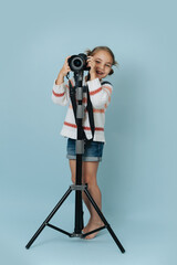 Cheerful little girl in striped sweater standing behind digital camera over blue