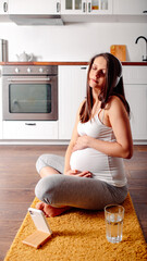 Pregnant doing workout on orange fitness mat. Pragnant yoga at home in the kitchen. Pregnant in white headphones.