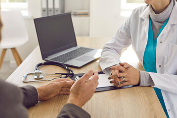 Close up of hands of unknown female doctor and male patient talk during medical consultation while sitting at table in hospital office. Healthcare and medicine concept. Selective focus.