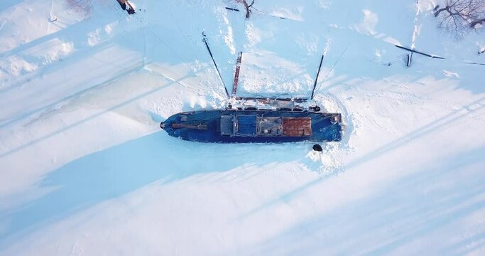 The drone rises above the ship with a blue deck, which is frozen into an icy river. All around is covered with white snow. Evening, sunny.