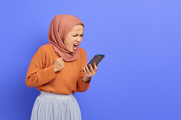 Angry young Asian woman in brown sweater and hijab using smartphone while loudly shouting isolated over purple background. People islam religious concept