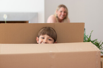 Funny little boy playing in new house. Schoolboy with dark hair hiding in big cardboard box. Mother in background. Real estate, purchase, family concept