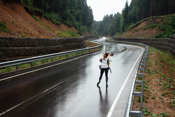 Young woman with yellow backpack walking on wet road among mountains. Female tourist spending leisure time for hiking on beautiful nature. Active lifestyles concept.