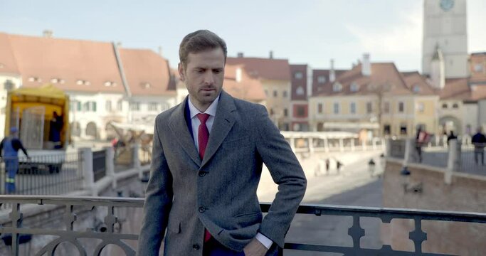 confident businessman in elegant suit wearing long coat, holding hand in pocket, looking to side, smiling when camera is zooming while standing outside in medieval city