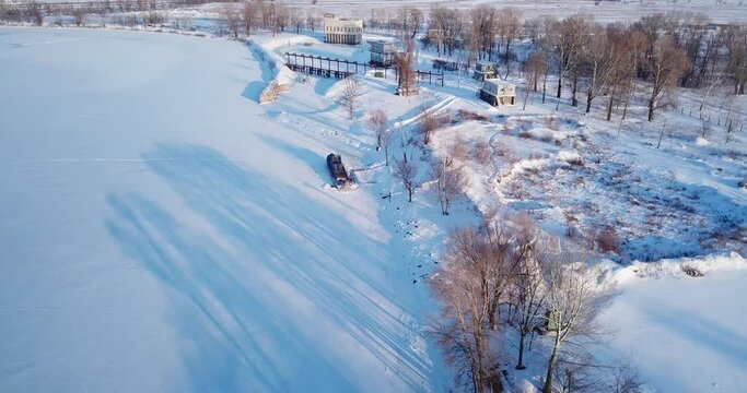 The drone flies along the bank of the river and flies up to a ship with a blue deck, which is frozen into an icy river. All around is covered with white snow. Evening, sunny.