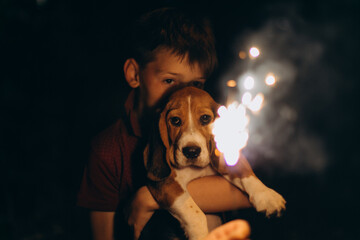 Caucasian boy and dog (beagle puppy) looking at bengal lights. Boy hugs his puppy. Evening event on fire background