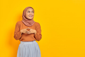 Beautiful smiling Asian woman in brown sweater and hijab holding hands together and feels optimistic,looking aside with confident isolated over purple background. People islam religious concept