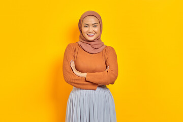 Beautiful smiling Asian woman in brown sweater and hijab crossed arms chest and looking confident isolated over yellow background. People islam religious concept