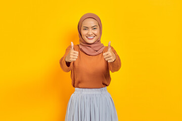 Beautiful smiling Asian woman in brown sweater and hijab showing thumbs up gesture with hand isolated over yellow background. People islam religious concept