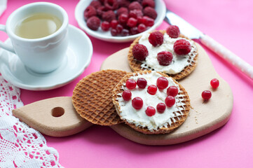 Waffles with cream cheese, cranberries and raspberries on a wooden board on a pink background. Romantic breakfast with fruits on waffles and cup of tea with white lace