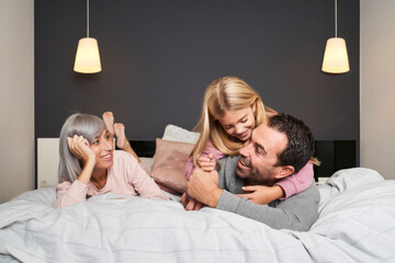 Family, consisting of father mothers and daughter sitting on the bed have fun tickling their...