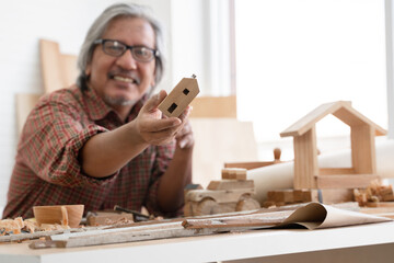 Selective focus on small wooden house model in Asian white haired senior carpenter man's hand. He smiling and showing wood work he made with pride at workplace at home. White background