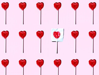Pattern of heart shaped lollipops flat laid against pink background with single one still in wrapping paper