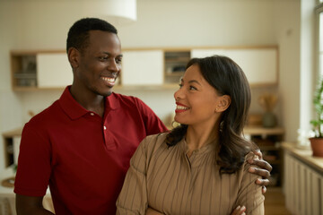 Handsome African American man in red polo t-shirt giving hug to his fiance, showing his tender attitude, love and passion, standing behind her, looking in eyes, cute dark-skinned woman smiling back