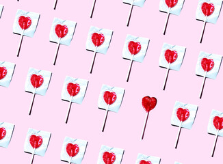 Pattern of heart shaped lollipops flat laid against pink background with single one unwrapped