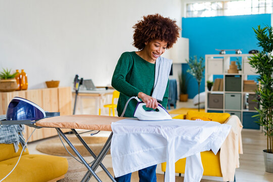 Smiling young Afro woman ironing shirt on board in living room