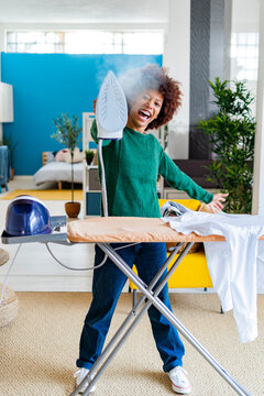 Cheerful young Afro woman blowing steam from iron standing by board with shirt in living room