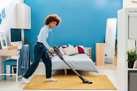 Cheerful Afro young woman cleaning carpet with vacuum cleaner in bedroom at home