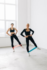 Fototapeta na wymiar Two smiling athletic Women are enjoying leg and buttocks work out with resistance band training together with friend in gym concept