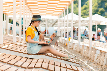 A young pretty woman is sitting on a chaise longue and typing on a laptop. Beach umbrellas in the...