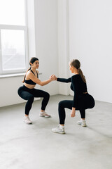Obraz premium Fitness pose two sporty women, blonde and brunette, in black sportswear show squats in studio, isolated at white, help each other holding hands - healthy lifestyle, training and friendship concept