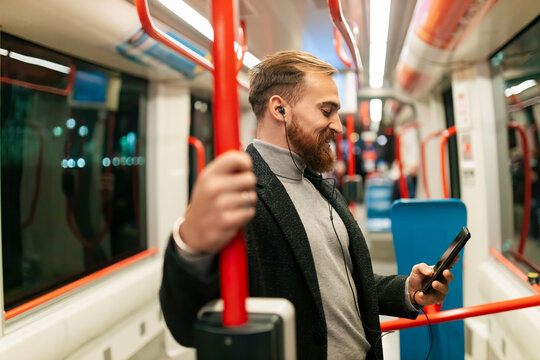 Man standing in tram using smart phone and listening music through in-ear headphones