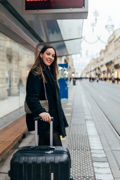 Smiling woman with suitcase waiting at tram station in city